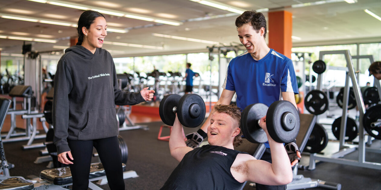 Male student holds dumb bells while a male and female student talk to him